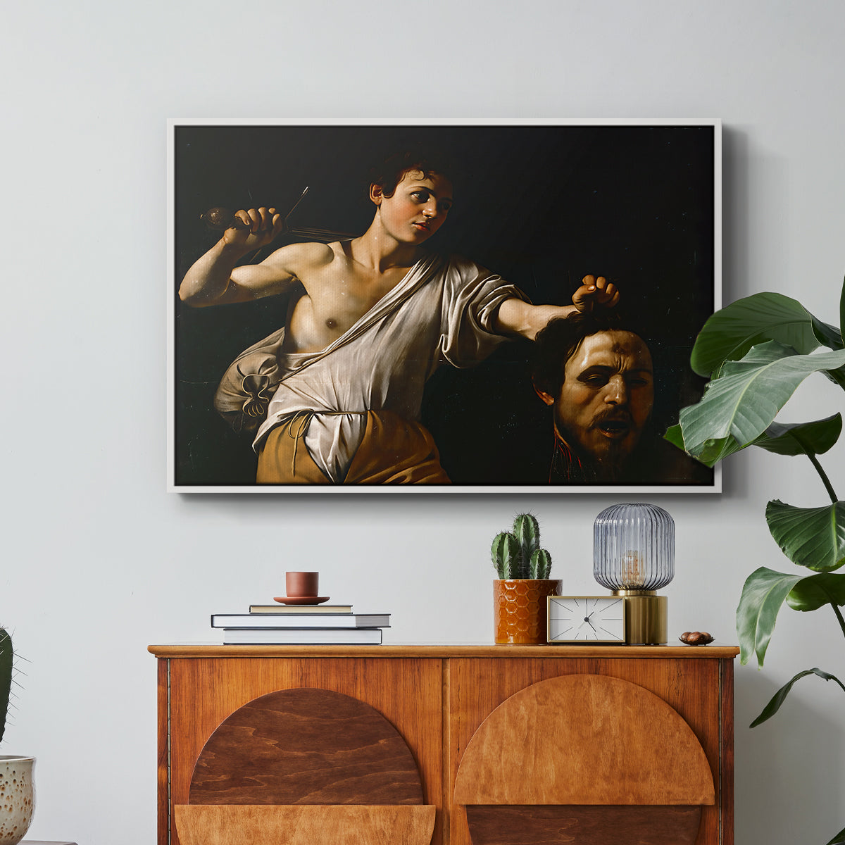 David with the Head of Goliath (Vienna Version), c. 1607 by Caravaggio in a white floater framed canvas featuring a young David holding the severed head of Goliath against a dark background, displayed above a wood console cabinet.
