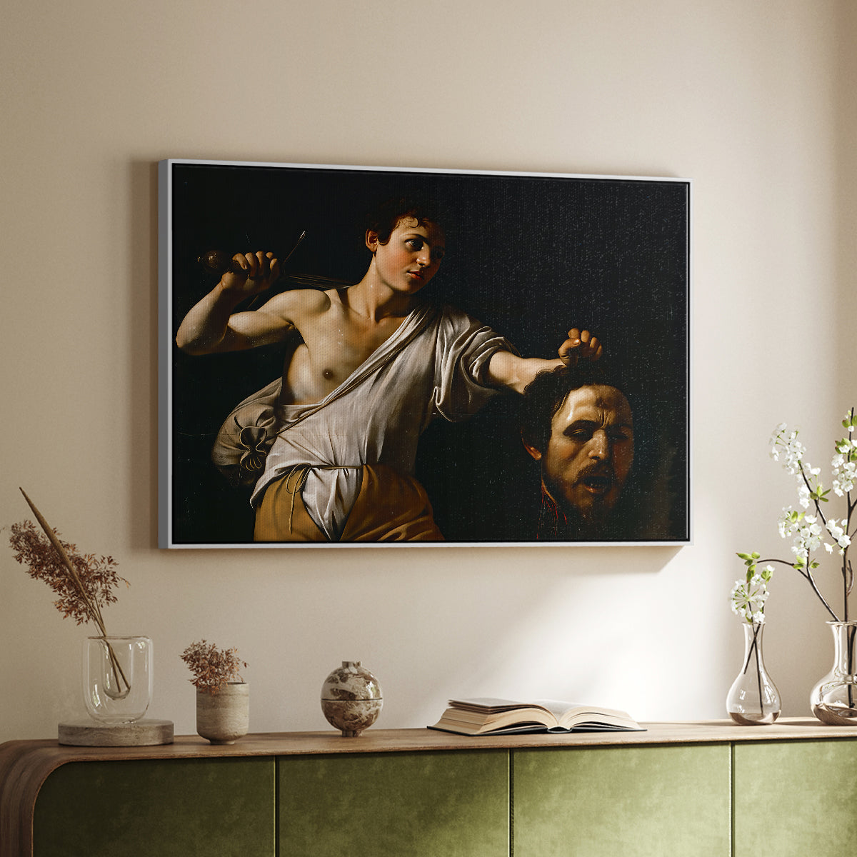 White floater framed canvas of David with the Head of Goliath (Vienna Version), c. 1607 by Caravaggio, featuring a biblical scene of David gripping Goliath's head with dramatic lighting above a sideboard in a softly lit hallway.