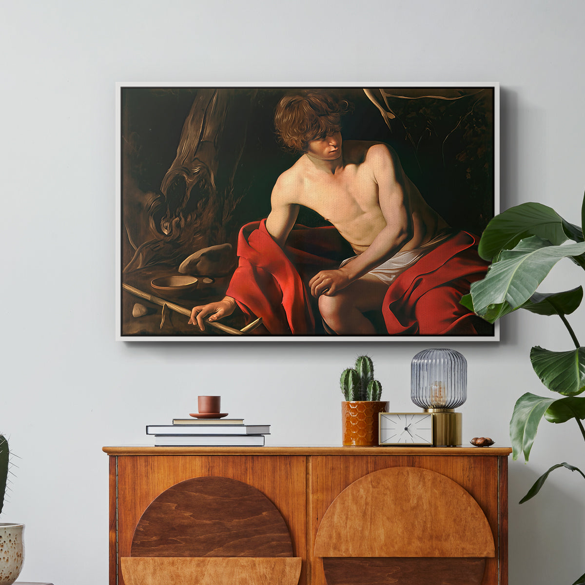 Saint John the Baptist in the Wilderness, c. 1603-1604 by Caravaggio in a white floater framed canvas featuring a young Saint John seated in wilderness with staff and red drapery, displayed above a wood console cabinet.