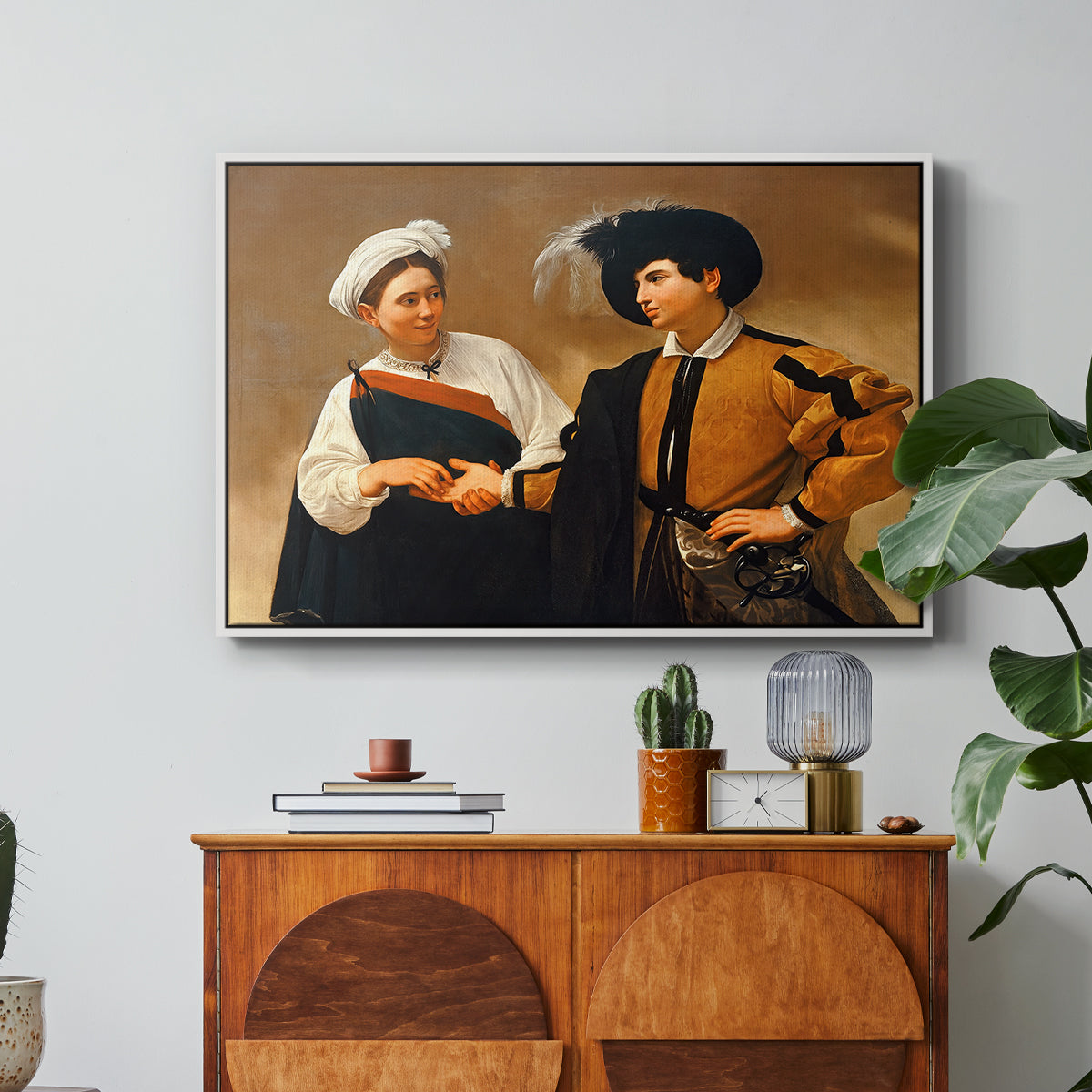 The Fortune Teller (Capitoline Version), c. 1599 by Caravaggio in a white floater framed canvas featuring a woman reading man's palm while discreetly removing his ring, displayed above a wood console cabinet.