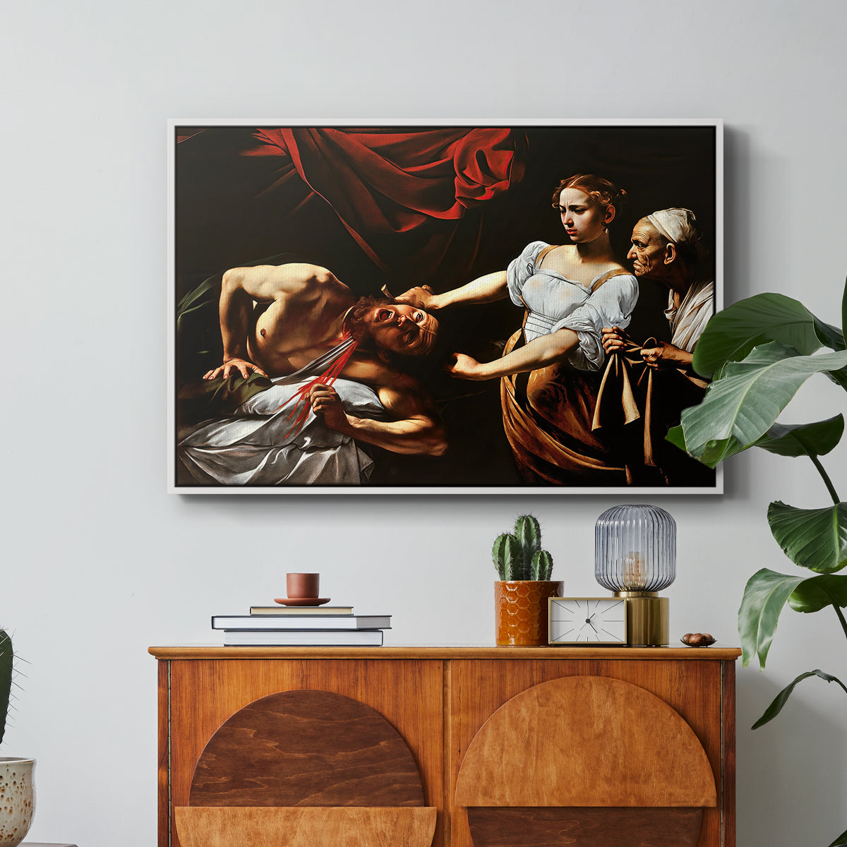 Judith Beheading Holofernes, c. 1599 by Caravaggio in a white floater framed canvas featuring Judith cutting off Holofernes' head as maidservant assists beside her, displayed above a wood console cabinet.