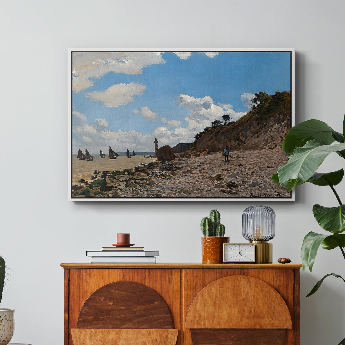 The Beach at Honfleur, 1864 by Claude Monet in a white floater framed canvas featuring a coastal beach scene with figures gathered along shoreline and boats nearby, displayed above a wood console cabinet.