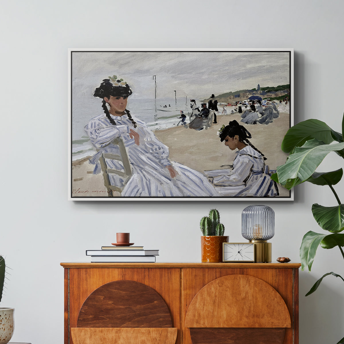 On the Beach at Trouville, 1870 by Claude Monet in a white floater framed canvas featuring figures seated on beach with parasols facing the ocean, displayed above a wood console cabinet.