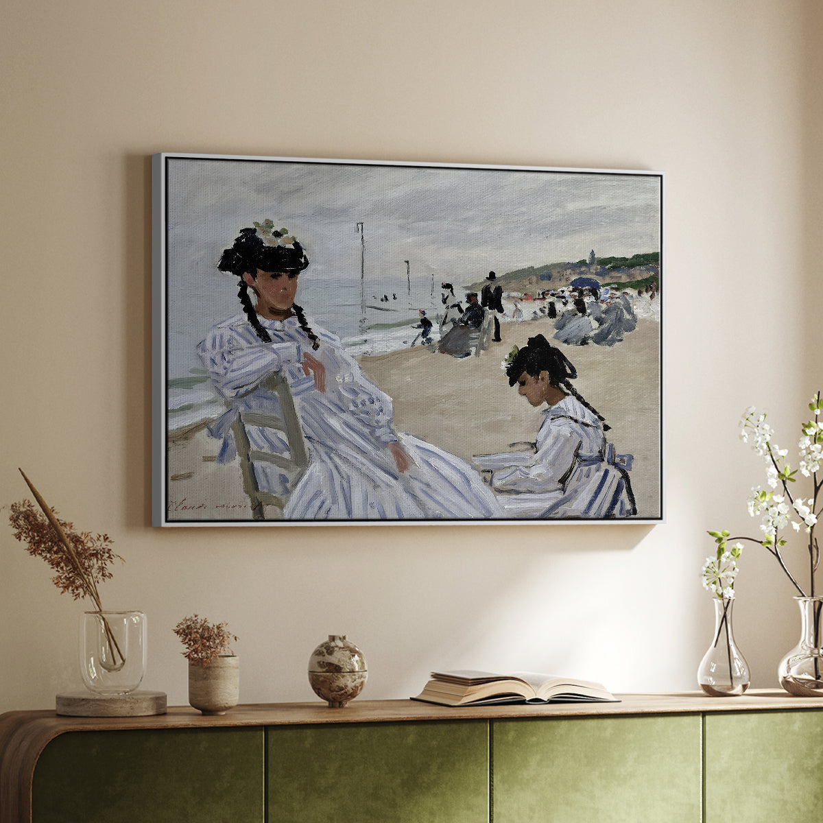 White floater framed canvas of On the Beach at Trouville, 1870 by Claude Monet, featuring an impressionist seaside scene with women in dresses and hats above a sideboard in a softly lit hallway.
