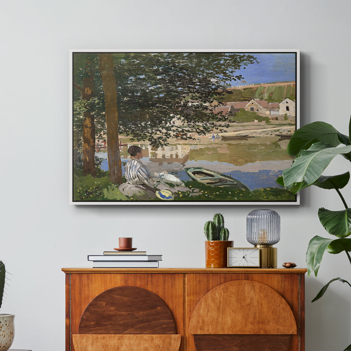On the Bank of the Seine, Bennecourt, 1868 by Claude Monet in a white floater framed canvas featuring a woman and child seated along riverbank beneath trees in soft daylight, displayed above a wood console cabinet.