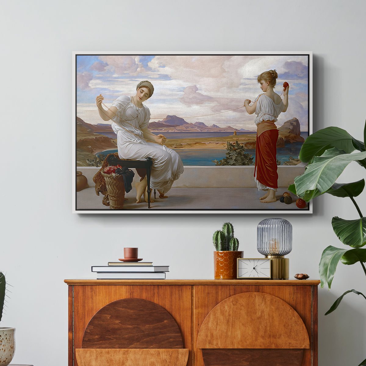 Winding the Skein, c. 1878 by Frederic Leighton in a white floater framed canvas featuring a woman holding thread while another figure helps wind yarn in classical setting, displayed above a wood console cabinet.