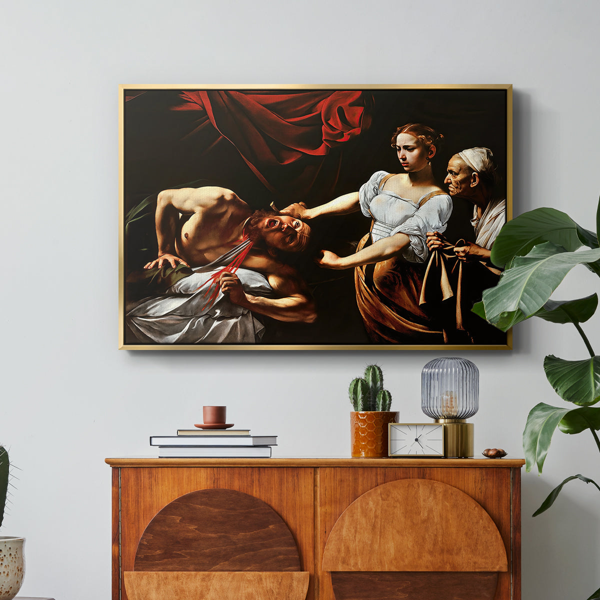 Judith Beheading Holofernes, c. 1599 by Caravaggio in a gold floater framed canvas featuring Judith cutting off Holofernes' head as maidservant assists beside her, displayed above a wood console cabinet.
