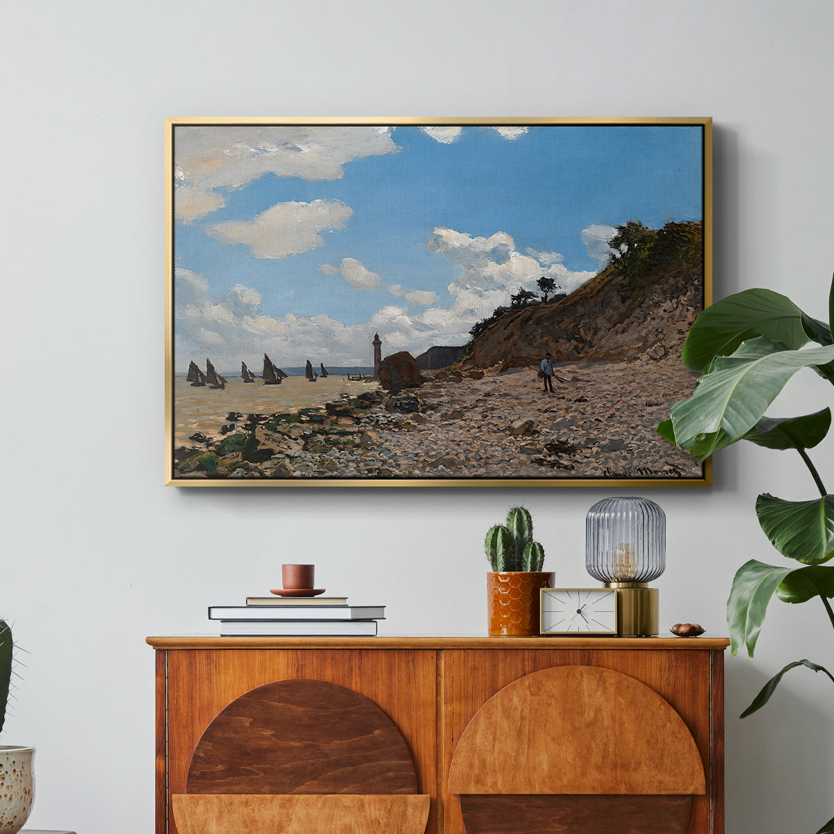 The Beach at Honfleur, 1864 by Claude Monet in a gold floater framed canvas featuring a coastal beach scene with figures gathered along shoreline and boats nearby, displayed above a wood console cabinet.