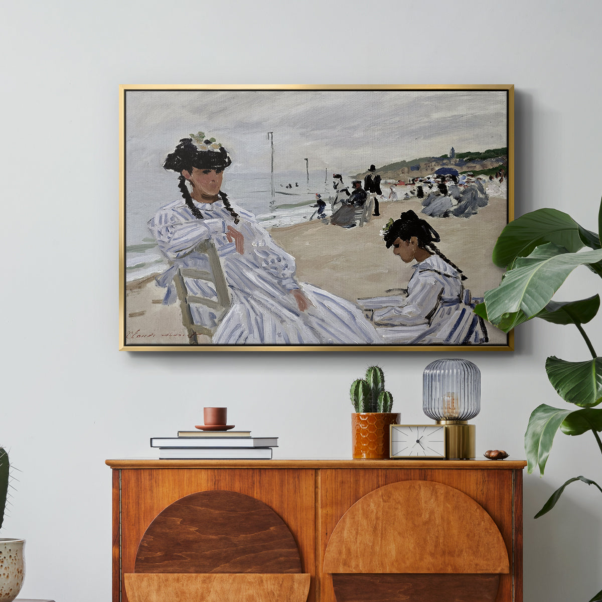 On the Beach at Trouville, 1870 by Claude Monet in a gold floater framed canvas featuring figures seated on beach with parasols facing the ocean, displayed above a wood console cabinet.