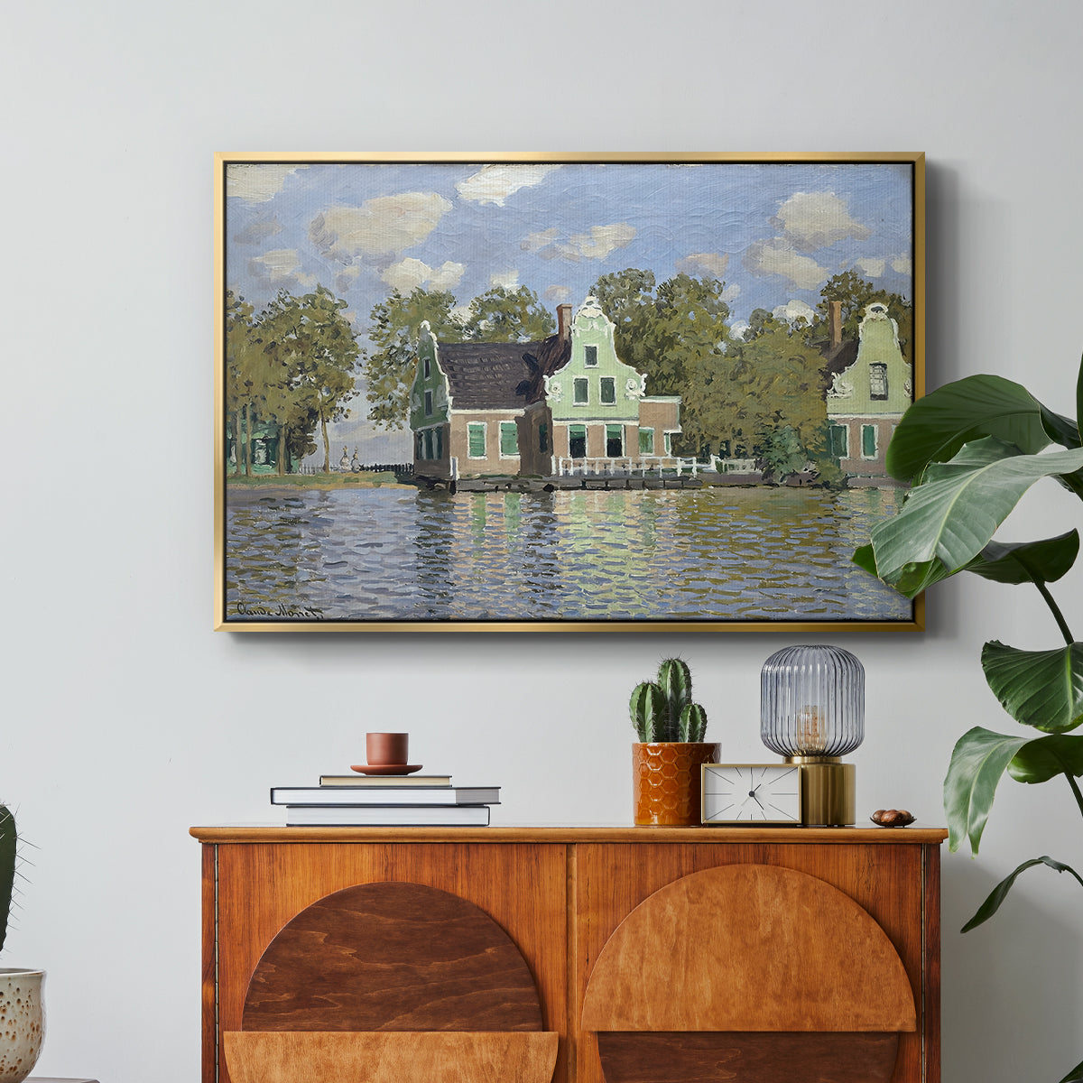 Houses on the Zaan River at Zaandam, 1871 by Claude Monet in a gold floater framed canvas featuring a row of houses along river with reflections in calm water, displayed above a wood console cabinet.