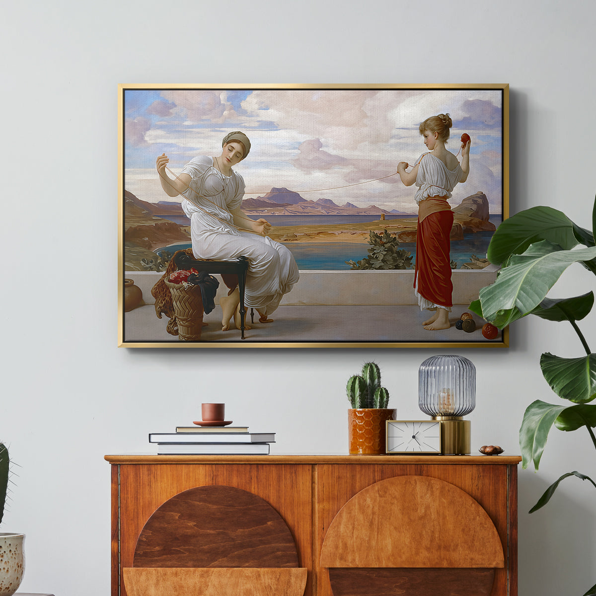 Winding the Skein, c. 1878 by Frederic Leighton in a gold floater framed canvas featuring a woman holding thread while another figure helps wind yarn in classical setting, displayed above a wood console cabinet.