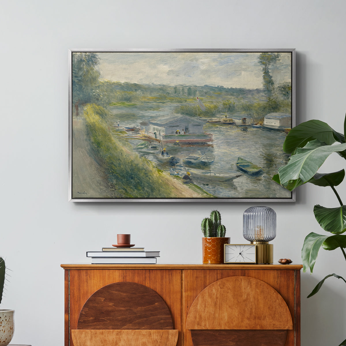 Wash-House Boat at Bas-Meudon, c. 1874 by Pierre-Auguste Renoir in a silver floater framed canvas featuring a floating wash-house boat along a riverbank with figures working nearby, displayed above a wood console cabinet.