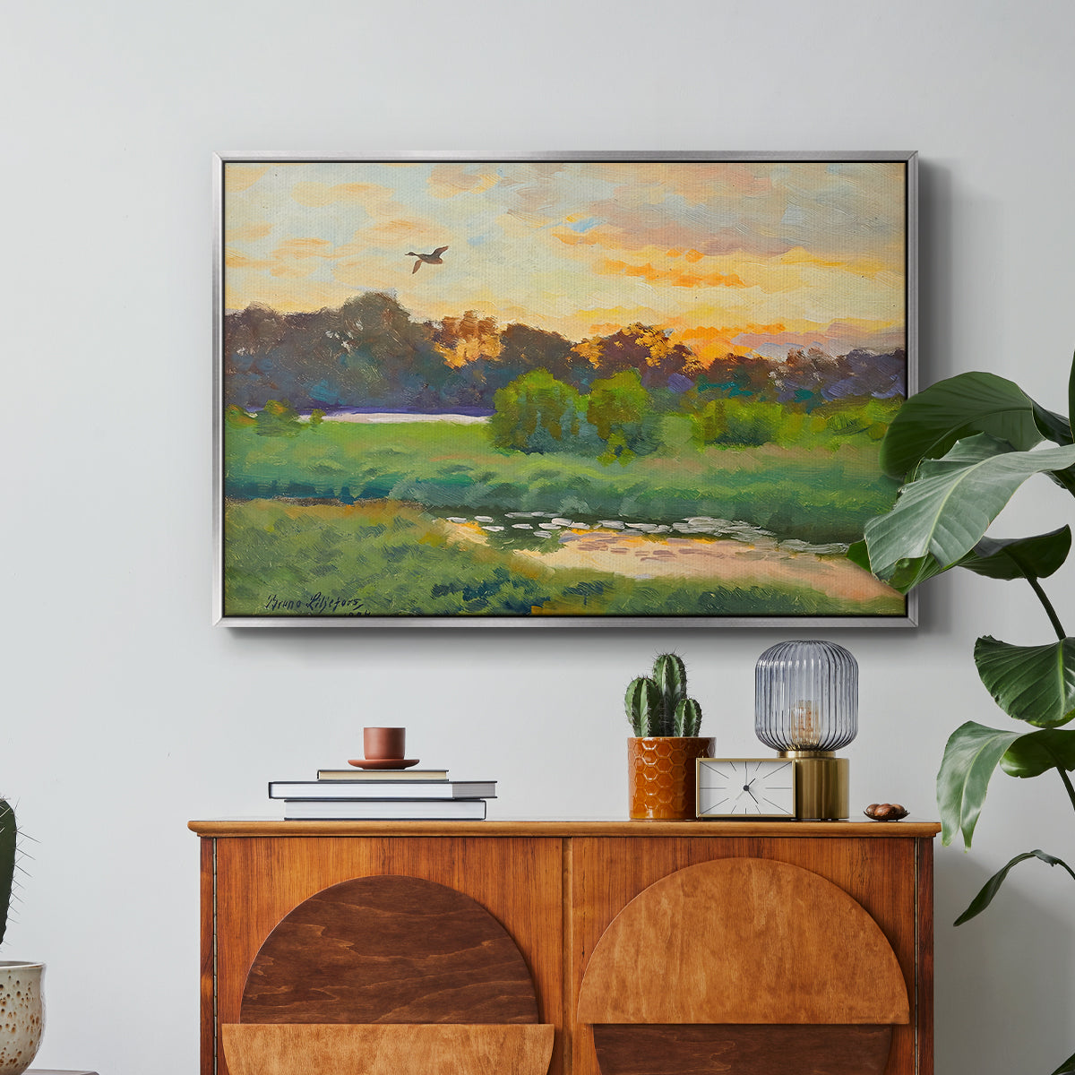 Flying Duck at Sunset, 1924 by Bruno Liljefors in a silver floater framed canvas featuring a wild duck flying over a sunset marsh with trees, displayed above a wood console cabinet.