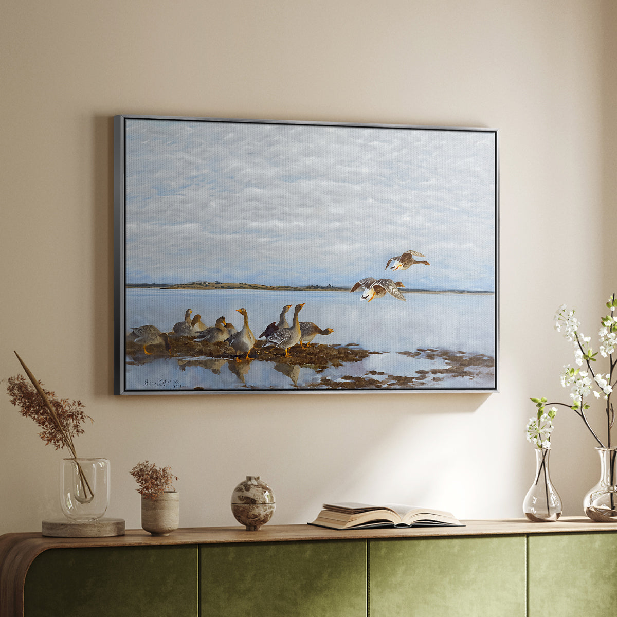 Silver floater framed canvas of Greylag Geese (Grågäss), 1922 by Bruno Liljefors, featuring birds standing and resting along shoreline with soft grass and water above a sideboard in a softly lit hallway.