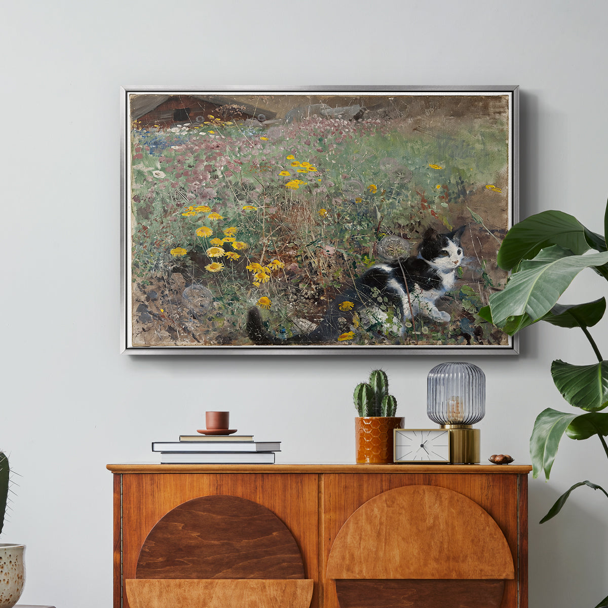 Cat on a Flowery Meadow, 1887 by Bruno Liljefors in a silver floater framed canvas featuring a cat crouching in a meadow filled with wildflowers and greenery, displayed above a wood console cabinet.
