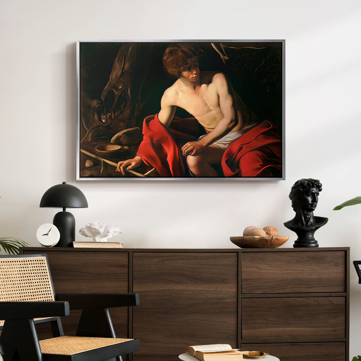 Saint John the Baptist in the Wilderness, c. 1603-1604 by Caravaggio displayed in a silver floater framed canvas above a dark wood credenza in an entryway.