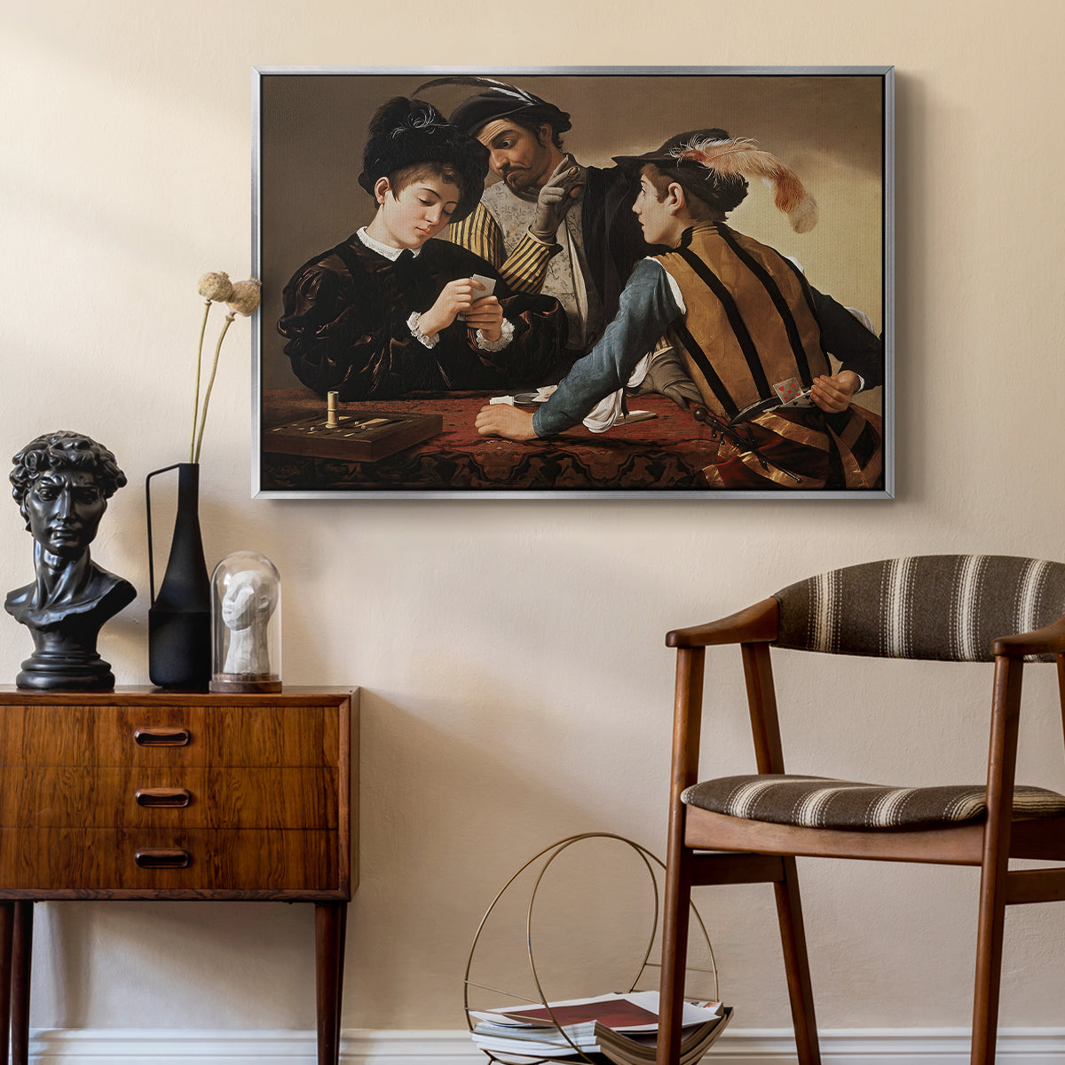 The Cardsharps, c. 1594 by Caravaggio as a silver floater framed canvas featuring a young men playing cards with one cheating and another watching closely, styled above a mid-century console in a contemporary living area.