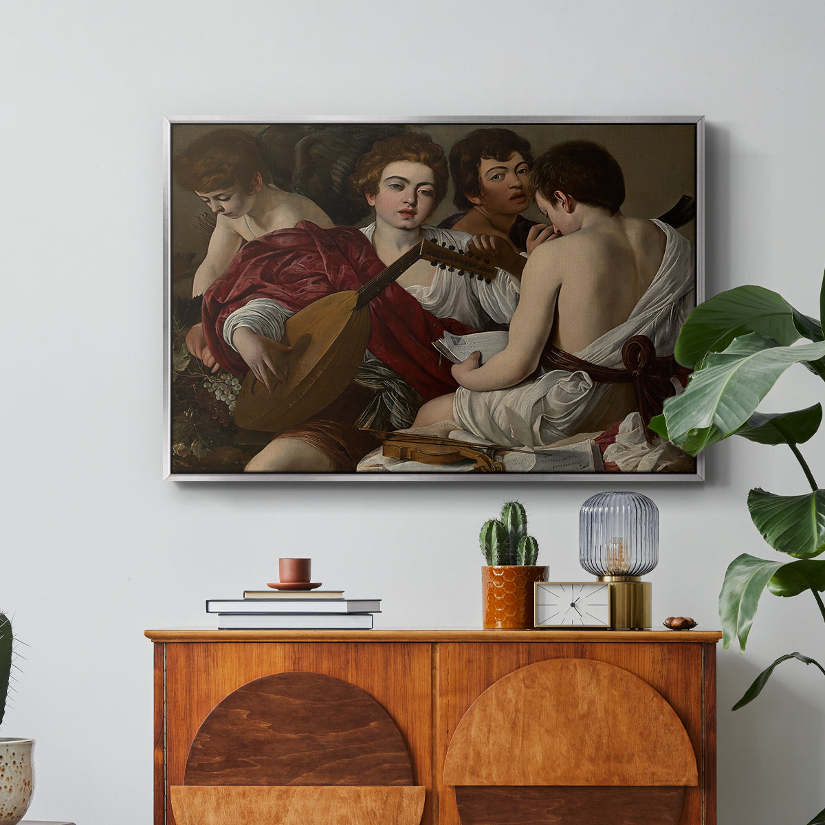 The Musicians, c. 1595 by Caravaggio in a silver floater framed canvas featuring a group of young men gathered together playing music with instruments, displayed above a wood console cabinet.