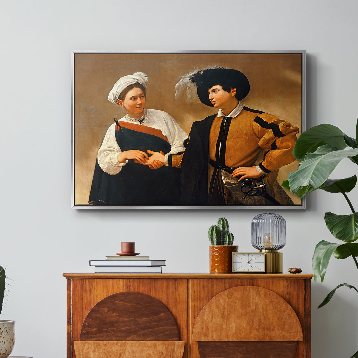 The Fortune Teller (Capitoline Version), c. 1599 by Caravaggio in a silver floater framed canvas featuring a woman reading man's palm while discreetly removing his ring, displayed above a wood console cabinet.