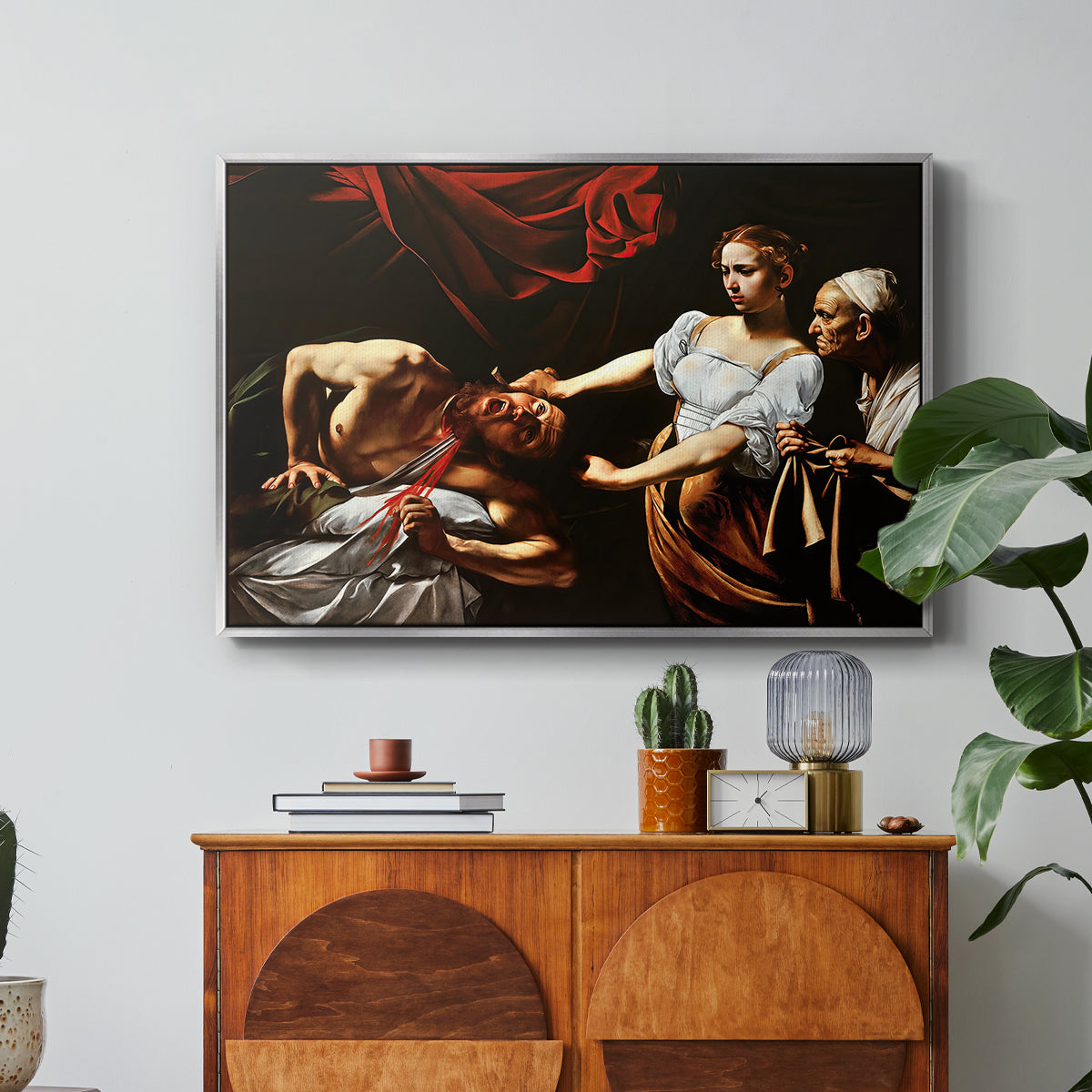 Judith Beheading Holofernes, c. 1599 by Caravaggio in a silver floater framed canvas featuring Judith cutting off Holofernes' head as maidservant assists beside her, displayed above a wood console cabinet.