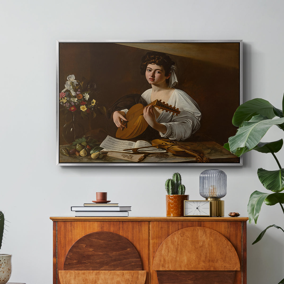 The Lute Player (Hermitage Version), c. 1596 by Caravaggio in a silver floater framed canvas featuring a young musician playing lute beside table with fruit and sheet music, displayed above a wood console cabinet.