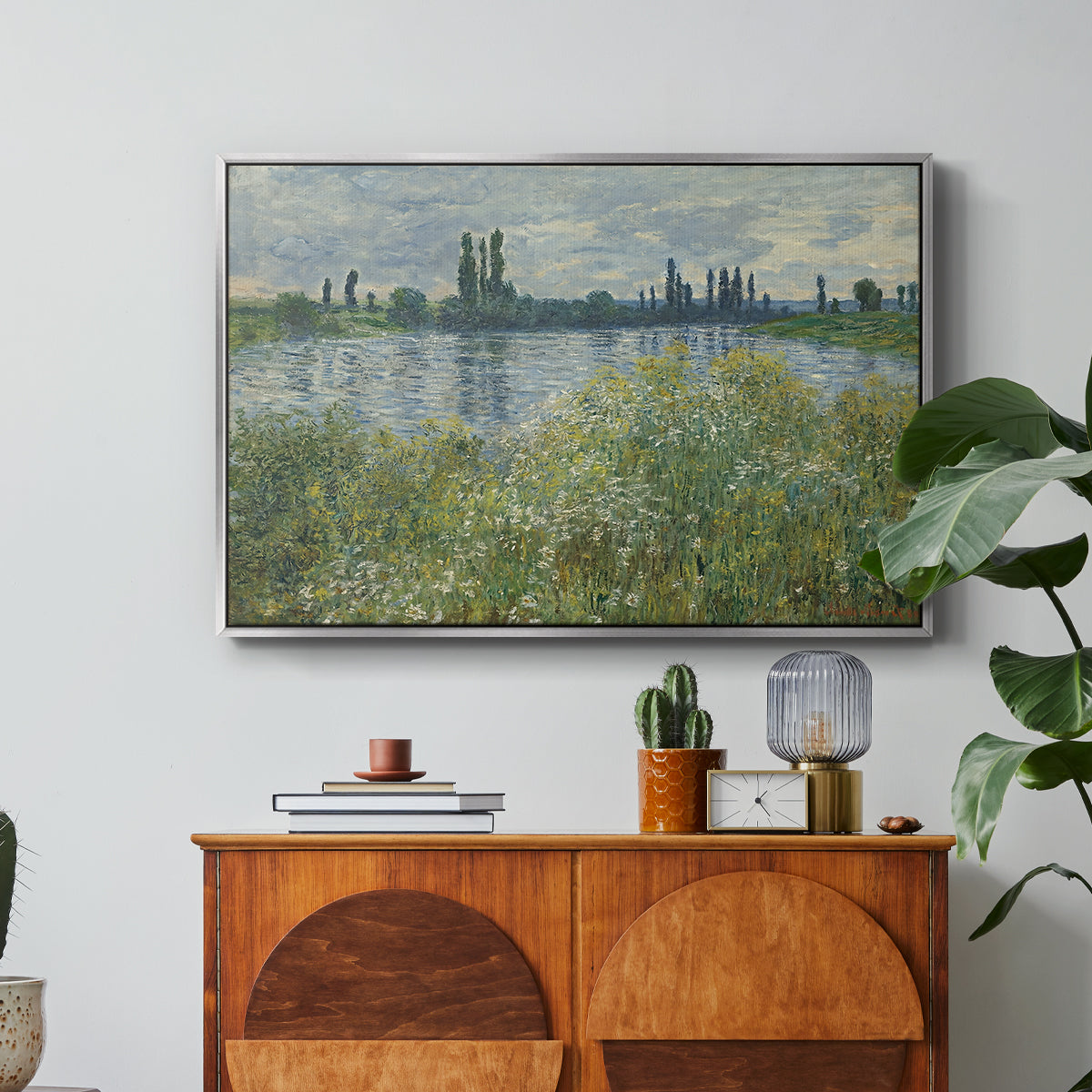 Banks of the Seine, Vétheuil, 1880 by Claude Monet in a silver floater framed canvas featuring a riverbank landscape with water, trees, and distant village buildings, displayed above a wood console cabinet.