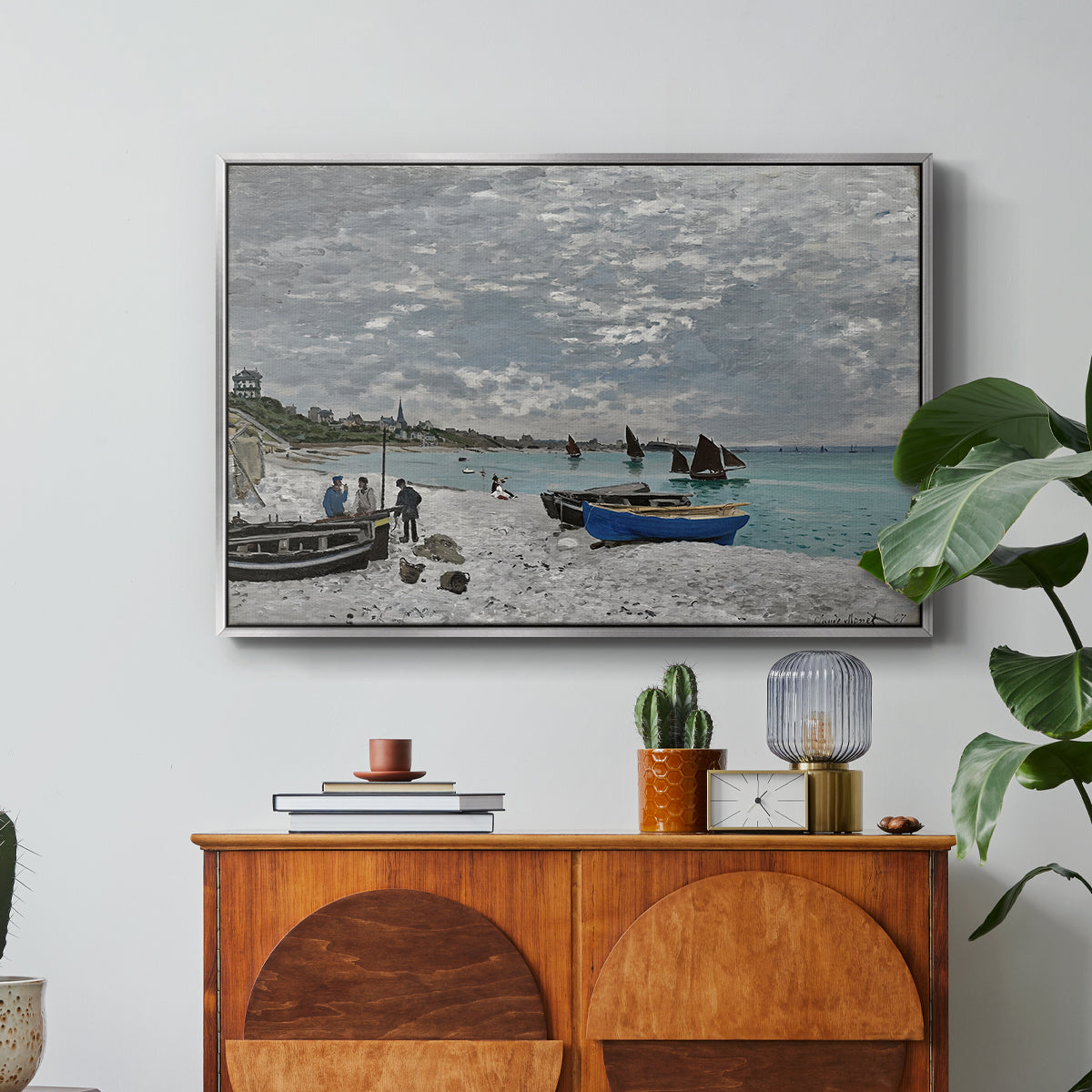 The Beach at Sainte-Adresse, 1867 by Claude Monet in a silver floater framed canvas featuring a seaside promenade with figures, flags, and boats along the coast, displayed above a wood console cabinet.