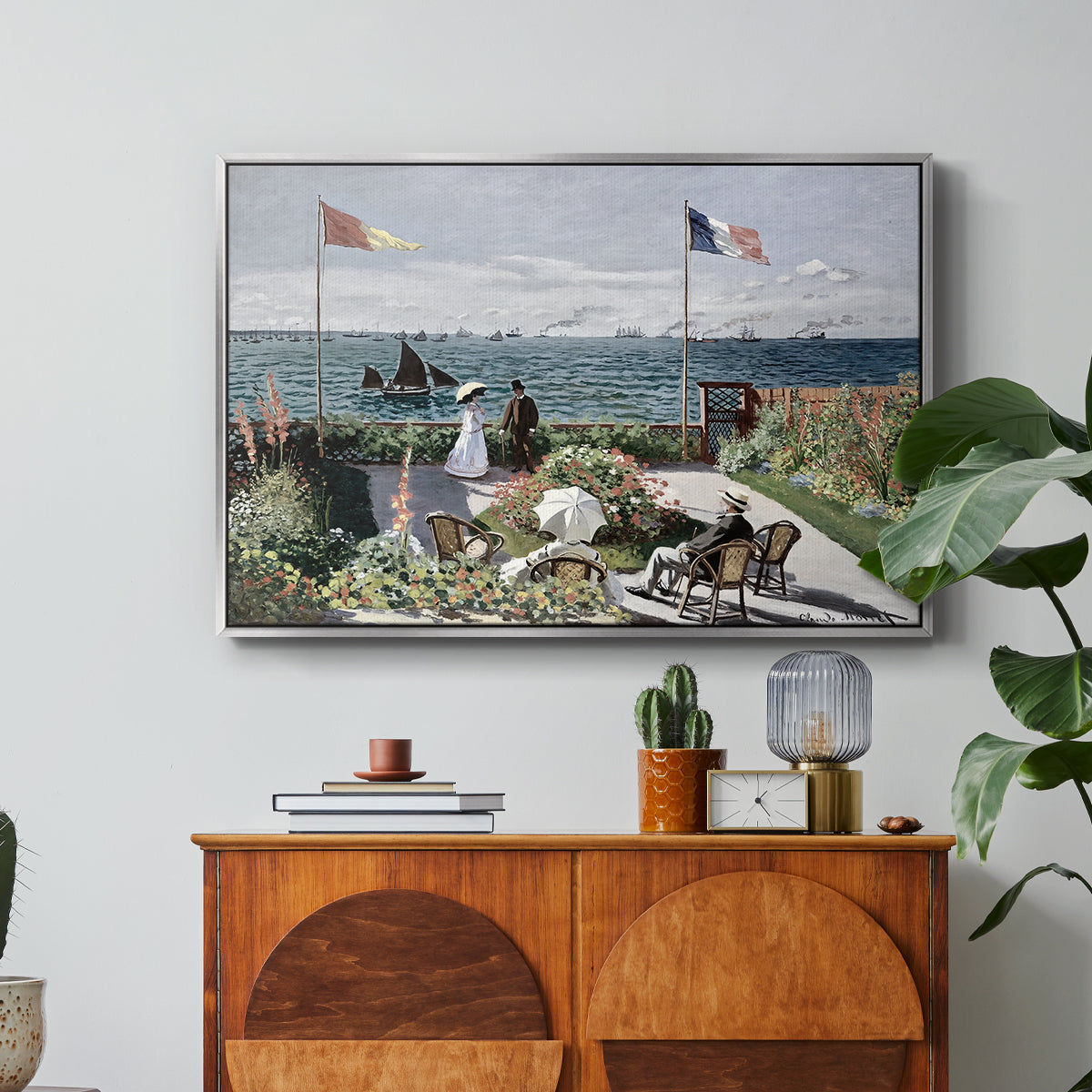 Garden at Sainte-Adresse, 1867 by Claude Monet in a silver floater framed canvas featuring a seaside garden with flowers, flags, and figures overlooking the water, displayed above a wood console cabinet.