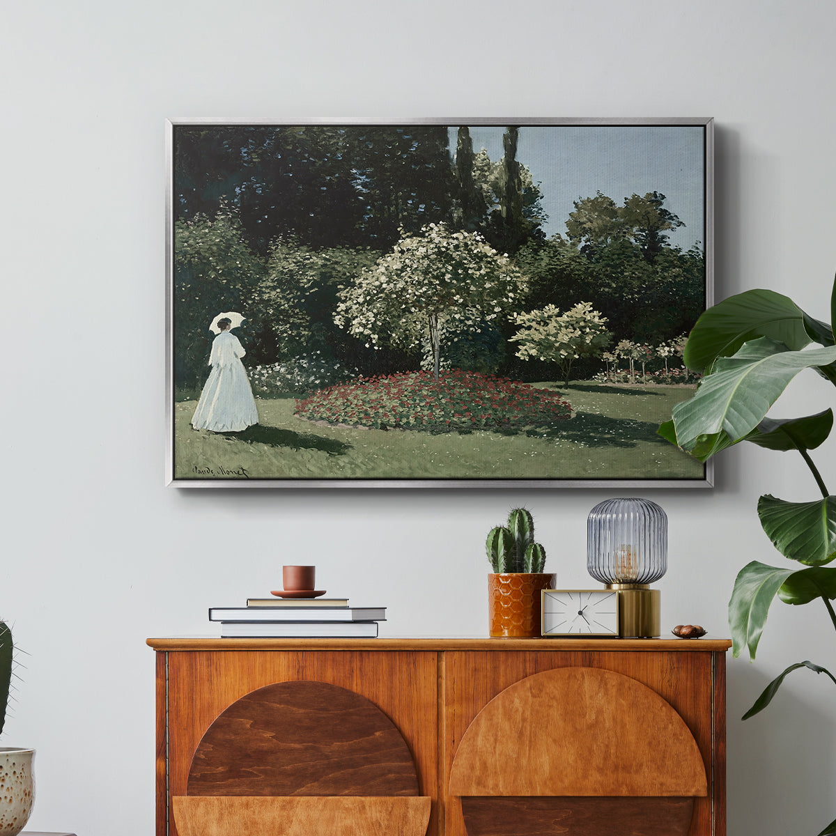 Woman in the Garden, Sainte-Adresse, 1867 by Claude Monet in a silver floater framed canvas featuring a woman standing in garden surrounded by flowers and greenery, displayed above a wood console cabinet.