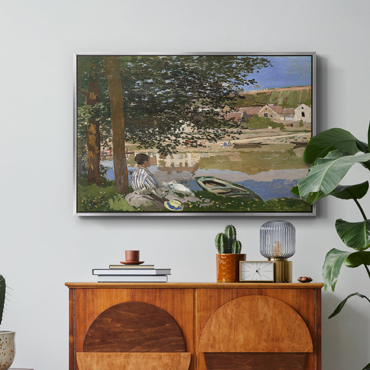 On the Bank of the Seine, Bennecourt, 1868 by Claude Monet in a silver floater framed canvas featuring a woman and child seated along riverbank beneath trees in soft daylight, displayed above a wood console cabinet.