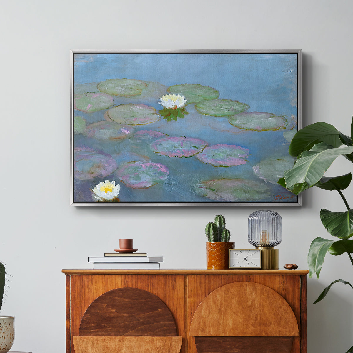 Water Lilies (Nymphéas), 1897-1899 by Claude Monet in a silver floater framed canvas featuring floating water lilies on pond with reflections of sky and foliage, displayed above a wood console cabinet.