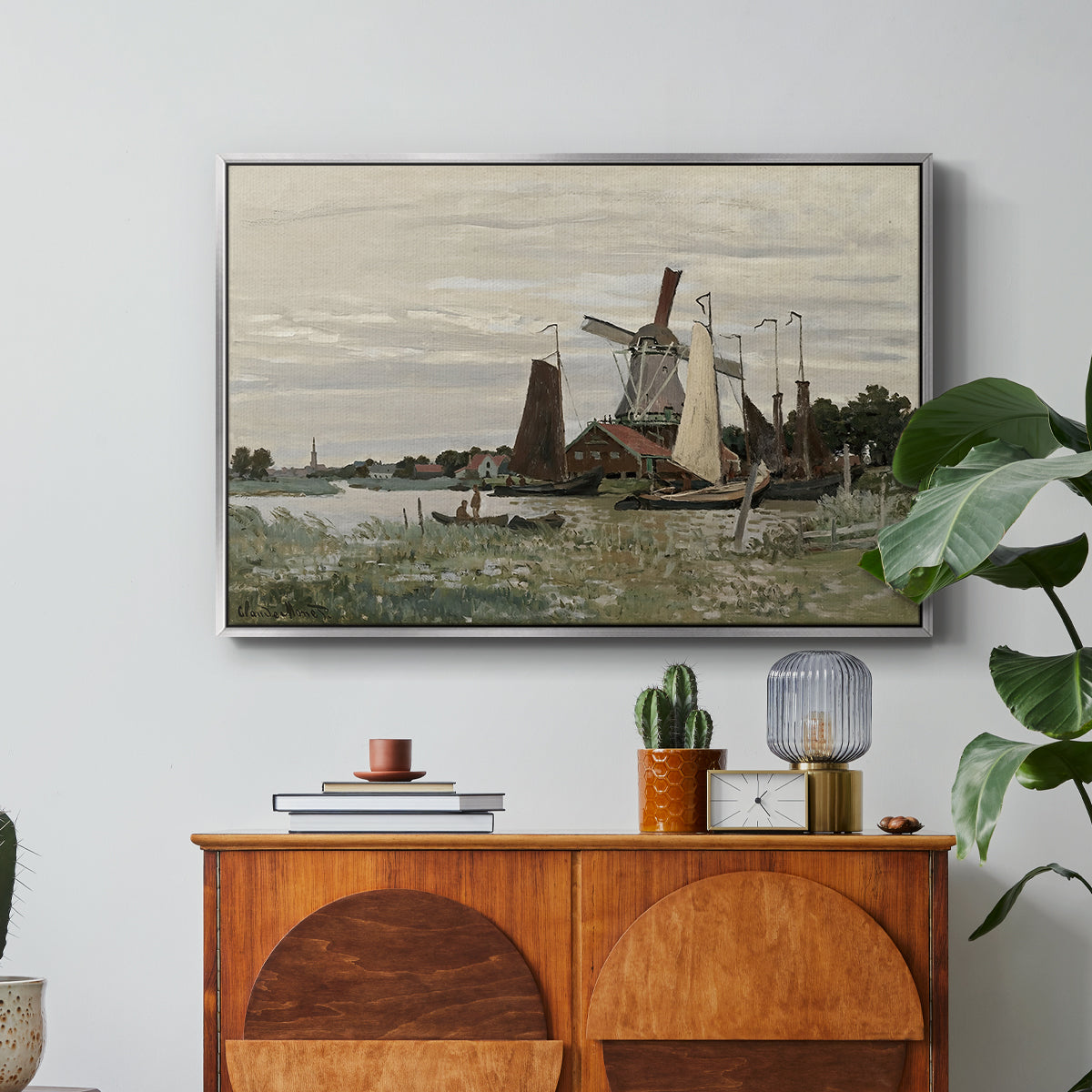 Windmill at Zaandam, 1871 by Claude Monet in a silver floater framed canvas featuring a single windmill beside water with surrounding buildings and sky, displayed above a wood console cabinet.