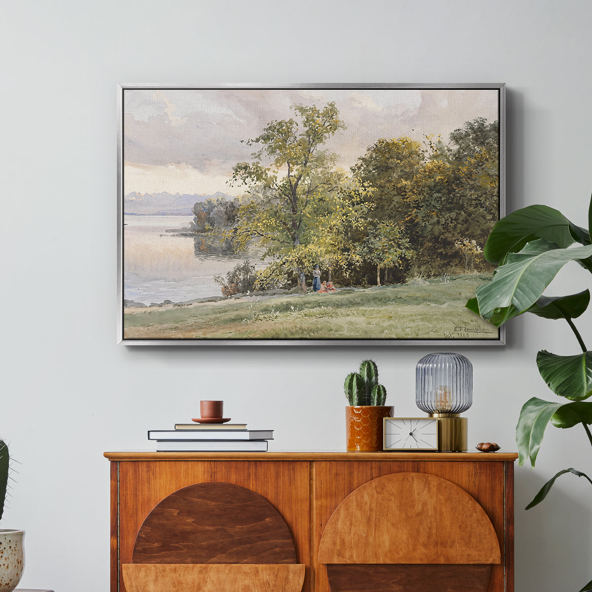 Landscape at Lake Starnberg, 1890 by Edward Theodore Compton in a silver floater framed canvas featuring a lakeside landscape with calm water and trees along shoreline, displayed above a wood console cabinet.