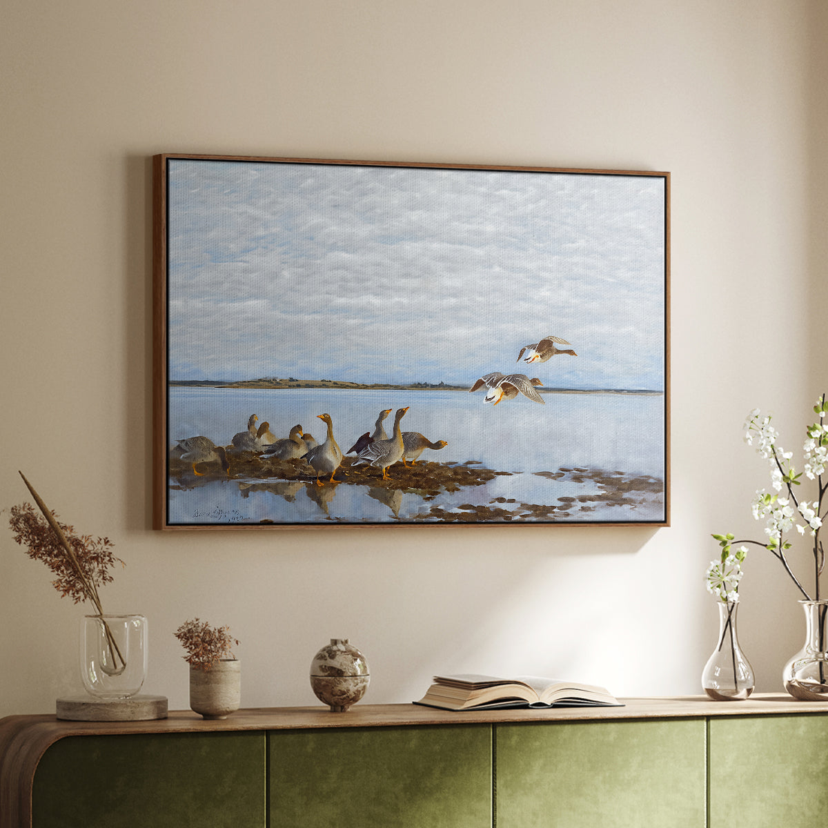 Walnut floater framed canvas of Greylag Geese (Grågäss), 1922 by Bruno Liljefors, featuring birds standing and resting along shoreline with soft grass and water above a sideboard in a softly lit hallway.