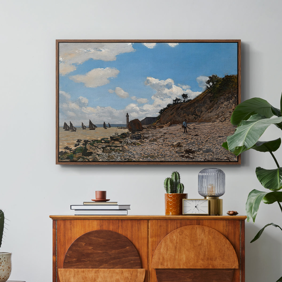 The Beach at Honfleur, 1864 by Claude Monet in a walnut floater framed canvas featuring a coastal beach scene with figures gathered along shoreline and boats nearby, displayed above a wood console cabinet.