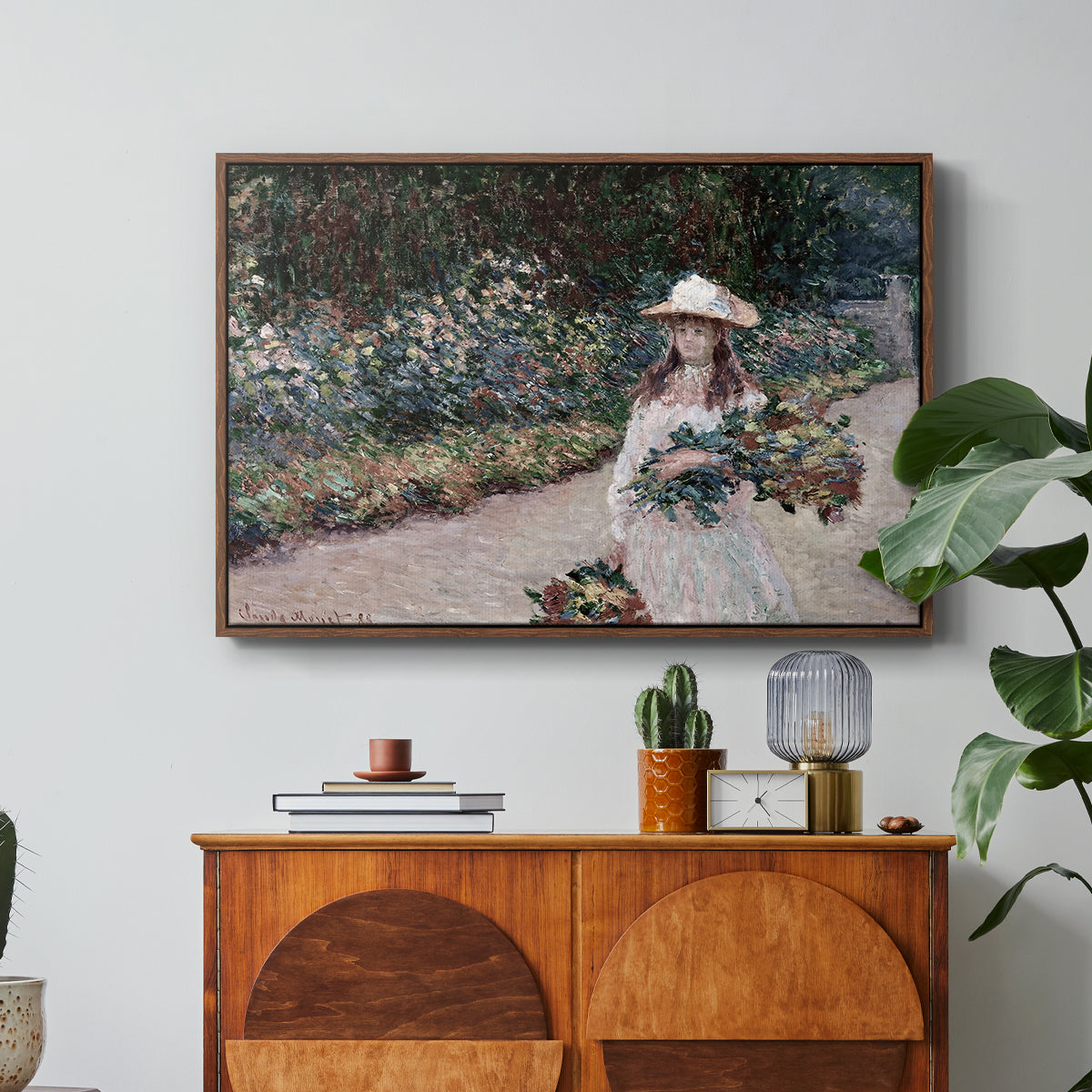 Young Girl in the Garden at Giverny, 1888 by Claude Monet in a walnut floater framed canvas featuring a young girl seated in lush garden surrounded by blooming flowers, displayed above a wood console cabinet.
