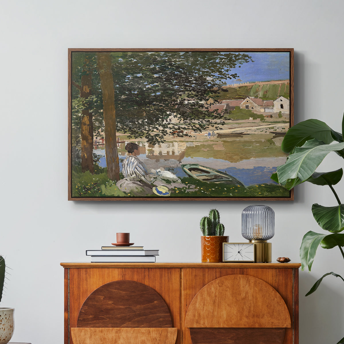 On the Bank of the Seine, Bennecourt, 1868 by Claude Monet in a walnut floater framed canvas featuring a woman and child seated along riverbank beneath trees in soft daylight, displayed above a wood console cabinet.