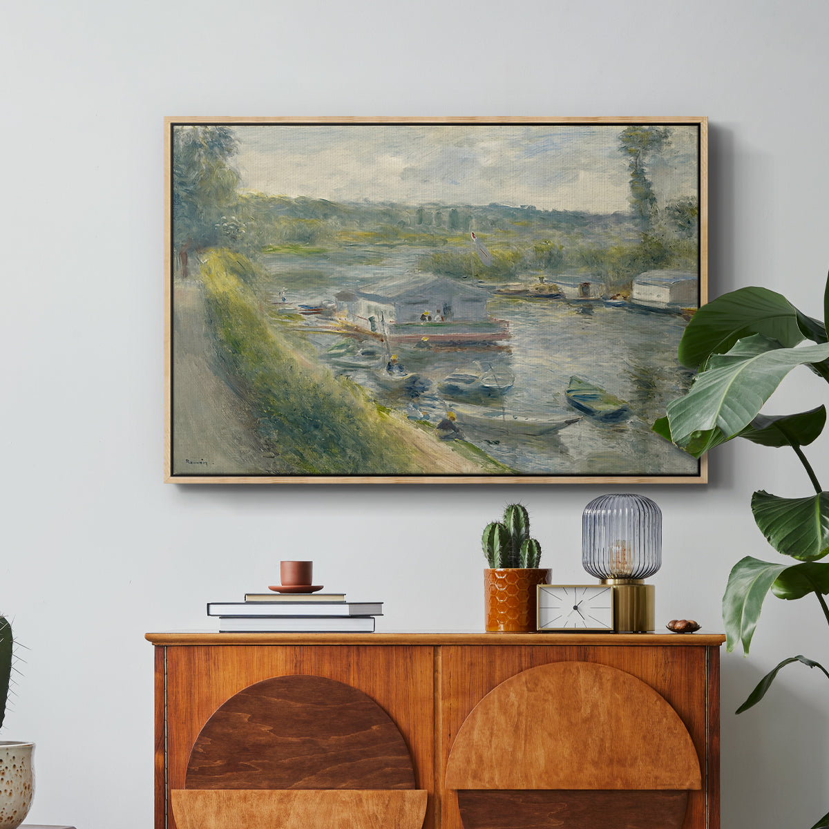 Wash-House Boat at Bas-Meudon, c. 1874 by Pierre-Auguste Renoir in a natural wood floater framed canvas featuring a floating wash-house boat along a riverbank with figures working nearby, displayed above a wood console cabinet.