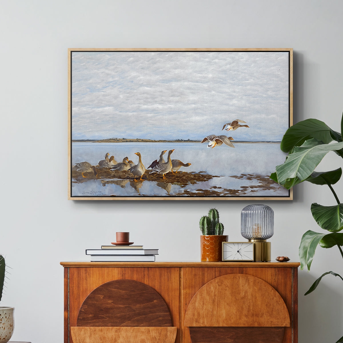 Greylag Geese (Grågäss), 1922 by Bruno Liljefors in a natural wood floater framed canvas featuring a group of geese gathered near water in a natural wetland landscape, displayed above a wood console cabinet.