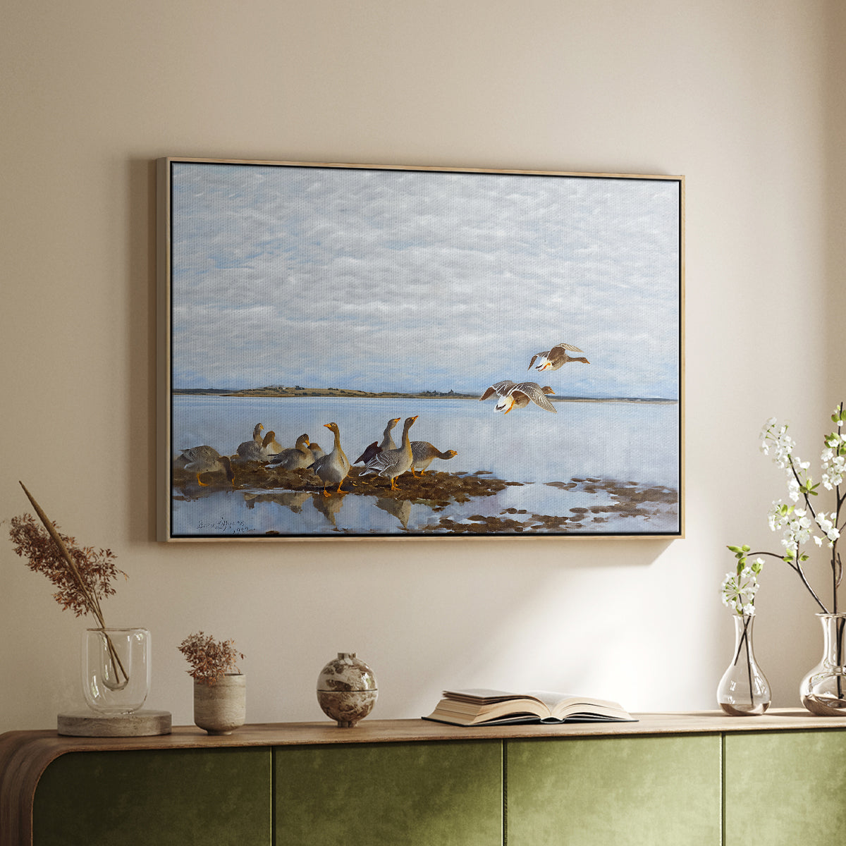 Natural wood floater framed canvas of Greylag Geese (Grågäss), 1922 by Bruno Liljefors, featuring birds standing and resting along shoreline with soft grass and water above a sideboard in a softly lit hallway.