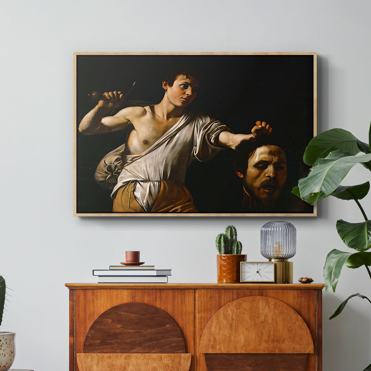David with the Head of Goliath (Vienna Version), c. 1607 by Caravaggio in a natural wood floater framed canvas featuring a young David holding the severed head of Goliath against a dark background, displayed above a wood console cabinet.