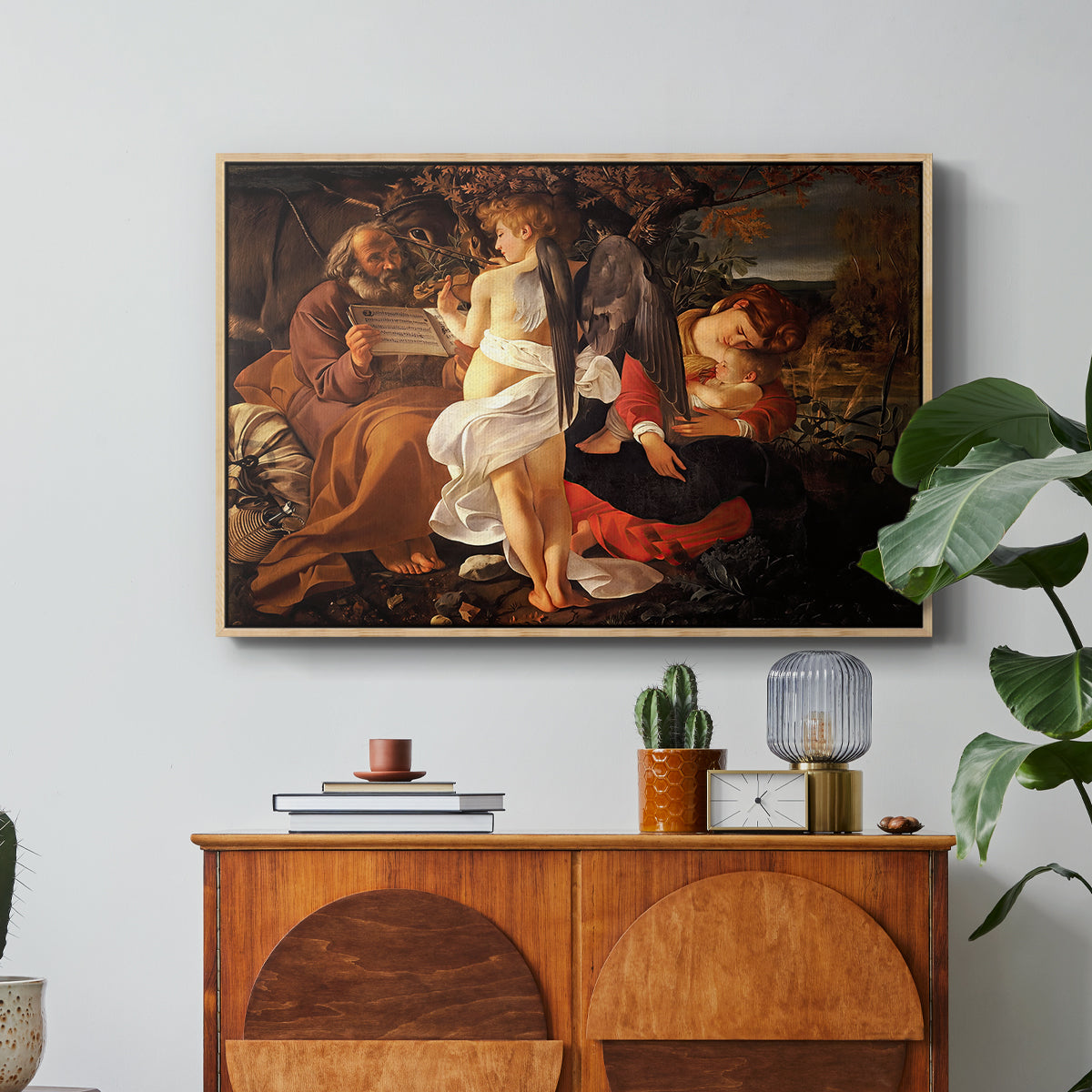 Rest on the Flight into Egypt, c. 1597 by Caravaggio in a natural wood floater framed canvas featuring the Holy Family resting in landscape with angel playing violin, displayed above a wood console cabinet.