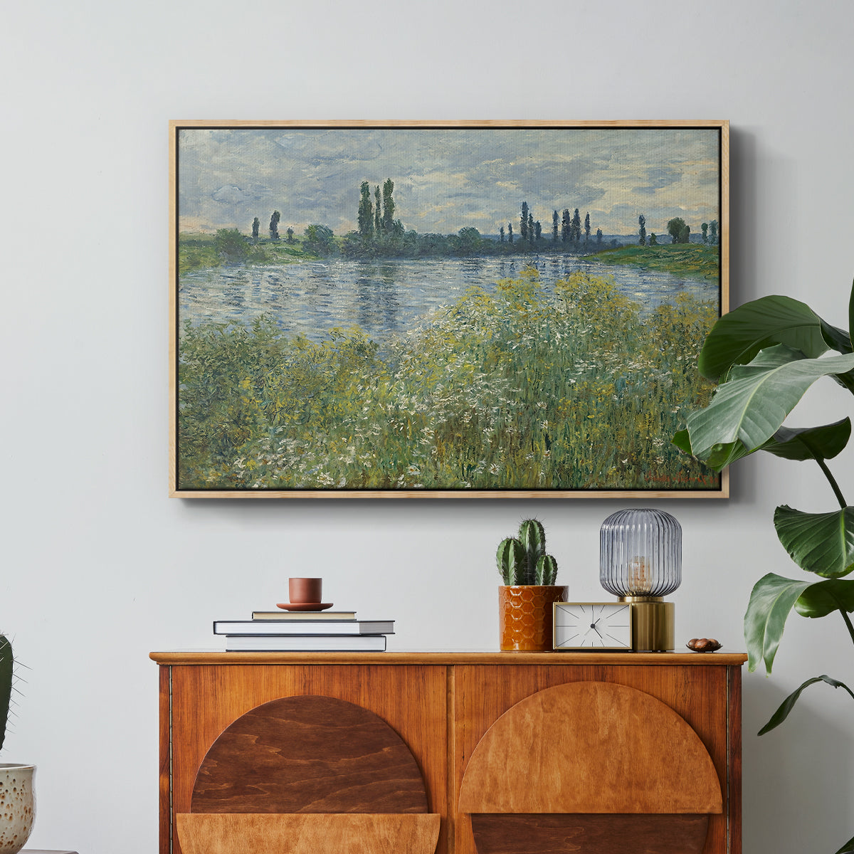 Banks of the Seine, Vétheuil, 1880 by Claude Monet in a natural wood floater framed canvas featuring a riverbank landscape with water, trees, and distant village buildings, displayed above a wood console cabinet.