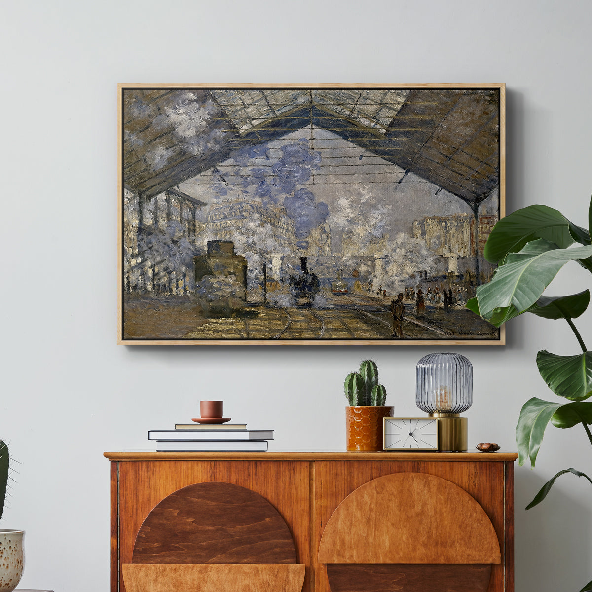 The Gare Saint-Lazare, 1877 by Claude Monet in a natural wood floater framed canvas featuring a train station interior filled with steam and locomotives under iron structure, displayed above a wood console cabinet.