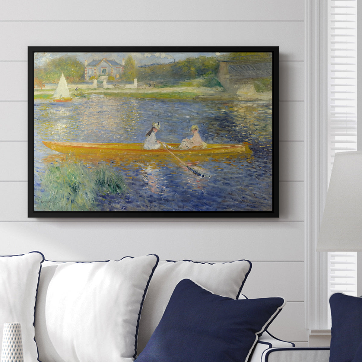 The Skiff (La Yole), 1875 by Pierre-Auguste Renoir displayed above a sofa in a living room in a black floater frame, featuring an impressionist boating scene with people relaxing in a narrow skiff.