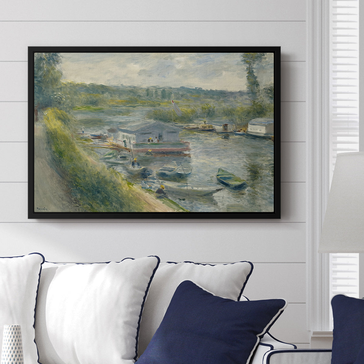 Wash-House Boat at Bas-Meudon, c. 1874 by Pierre-Auguste Renoir displayed above a sofa in a living room in a black floater frame, featuring a riverside scene with moored boat used for washing clothes.