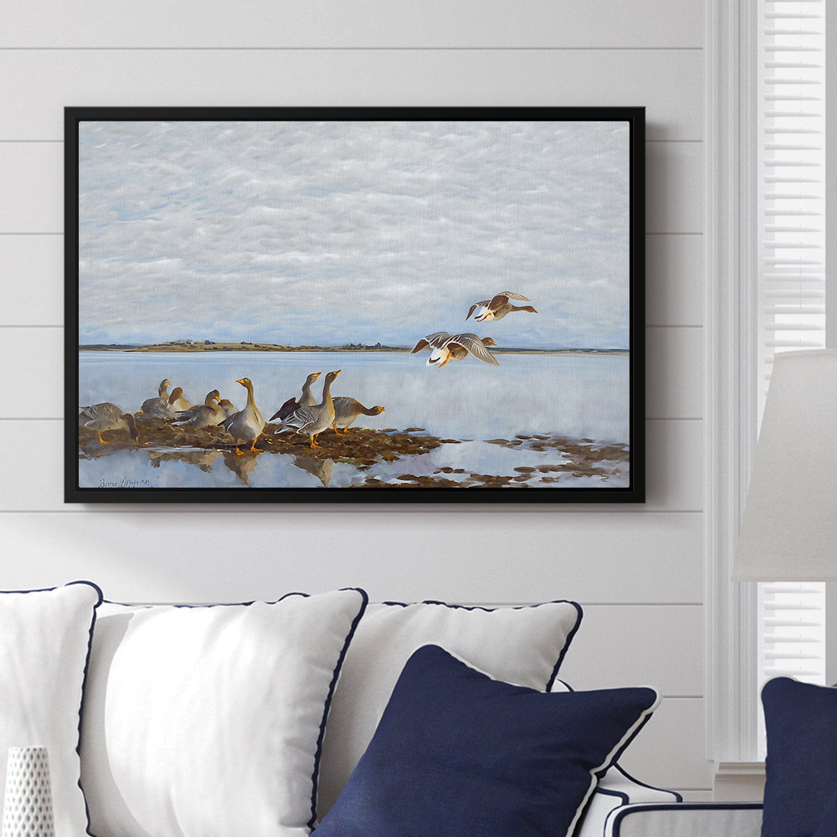 Greylag Geese (Grågäss), 1922 by Bruno Liljefors displayed above a sofa in a living room in a black floater frame, featuring birds standing and resting along shoreline with soft grass and water.