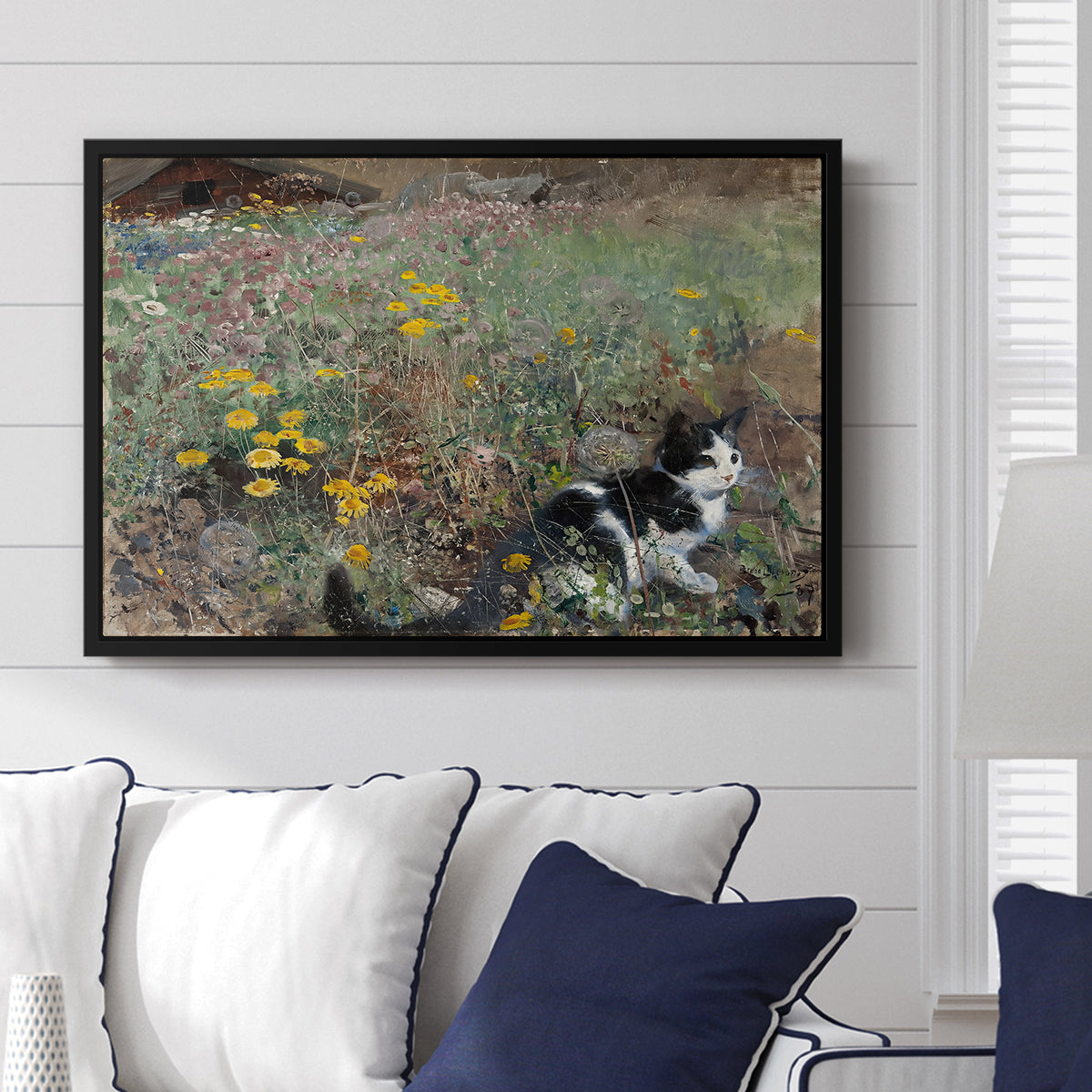 Cat on a Flowery Meadow, 1887 by Bruno Liljefors displayed above a sofa in a living room in a black floater frame, featuring a small cat resting among colorful flowers in grassy field.