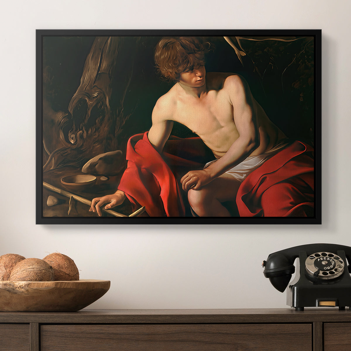 Saint John the Baptist in the Wilderness, c. 1603-1604 by Caravaggio in a black floater framed canvas displayed above a console table in a hallway, featuring a contemplative religious figure in rustic wilderness environment.
