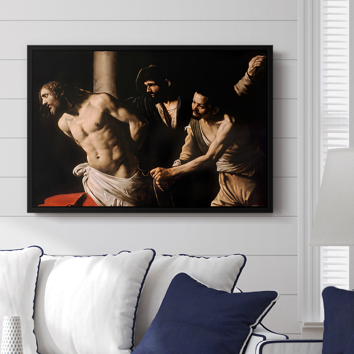 The Flagellation of Christ at the Column (Rouen Version), 1606 by Caravaggio displayed above a sofa in a living room in a black floater frame, featuring a dramatic religious scene of Christ enduring flagellation in dark interior.